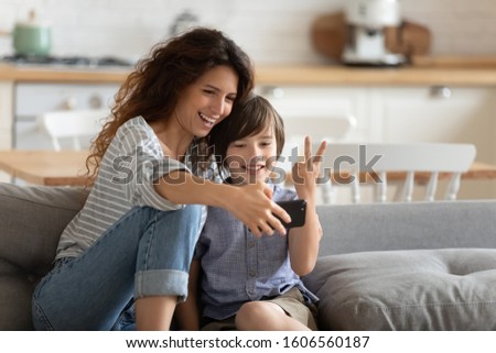 Cheerful mother holding smart phone making selfie with little school age son showing victory sign, mom and kid boy sit on couch using gadget having fun taking self-portrait spend time together at home