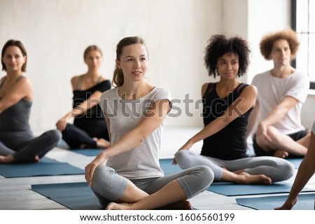 Sporty young diverse students practicing yoga exercise at group lesson, sitting in Parivritta Sukhasana pose on mats, Caucasian and African American people working out in modern fitness center Royalty-Free Stock Photo #1606560199