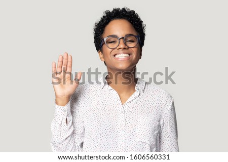Head shot portrait african pretty young woman in glasses smiles looks at camera raise palm wave hand makes gesture of greeting friendly attitude good relation posing isolated on grey studio background Royalty-Free Stock Photo #1606560331