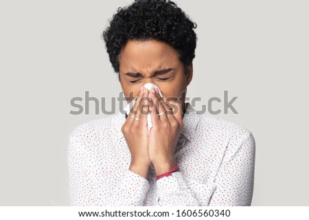 Allergic ill young african woman holding tissue blowing running nose got chill sneezing in handkerchief feels unwell, head shot portrait isolated on grey background, influenza virus snuffles concept Royalty-Free Stock Photo #1606560340
