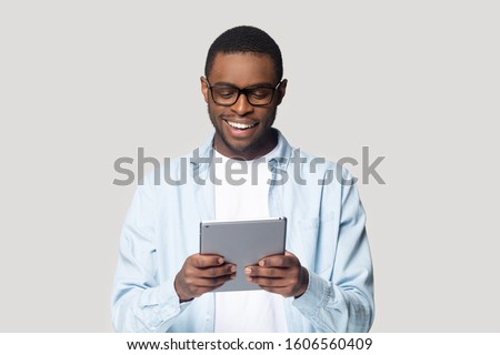 Smiling african guy hold tablet digital device having fun using application browsing sites, shopping in internet, flirting with girlfriend via online dating websites isolated on gray studio background Royalty-Free Stock Photo #1606560409