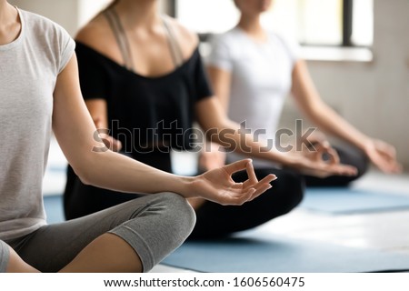 Young woman practicing yoga at group lesson, doing Sukhasana exercise, people sitting in Easy Seat pose, mudra hands close up, stress relief and wellbeing, working out in modern yoga center club Royalty-Free Stock Photo #1606560475