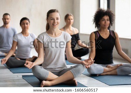 Smiling young woman enjoying practicing yoga at group lesson, sitting in Easy Seat pose on mat, diverse people doing Sukhasana exercise, stress relief, working out in modern yoga center club Royalty-Free Stock Photo #1606560517
