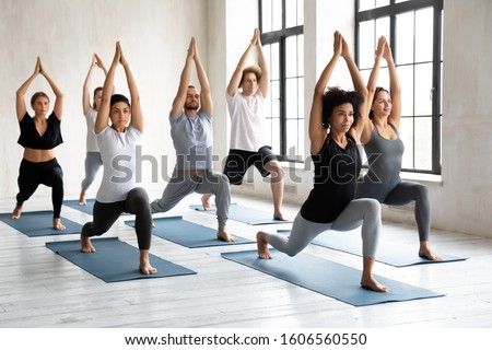 Young diverse young people with African American instructor doing Warrior one exercise, practicing yoga at group lesson, standing in Virabhadrasana pose, working out in modern yoga studio Royalty-Free Stock Photo #1606560550