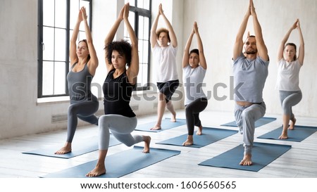 Diverse young sporty people doing Warrior one exercise at group lesson, practicing yoga in modern fitness center, standing in Virabhadrasana pose, working out with African American female instructor Royalty-Free Stock Photo #1606560565