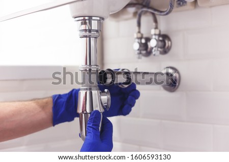 Plumber repairs and maintains chrome siphon under the washbasin. Plumber at work in bathroom, plumbing assemble and install concept Royalty-Free Stock Photo #1606593130