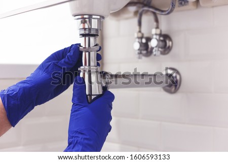 Plumber repairs and maintains chrome siphon under the washbasin. Plumber at work in bathroom, plumbing assemble and install concept Royalty-Free Stock Photo #1606593133