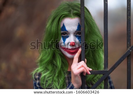 Close-up portrait of a greenhaired girl in chekered dress with joker makeup on a blurry brown background.