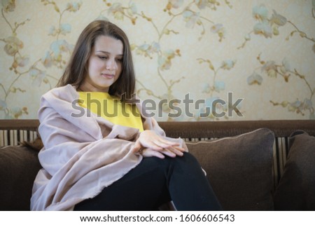 A picture of nice and thoughtful girl sit at sofa and reads a book. She has brown blanket on her shoulders. Woman got cold. 