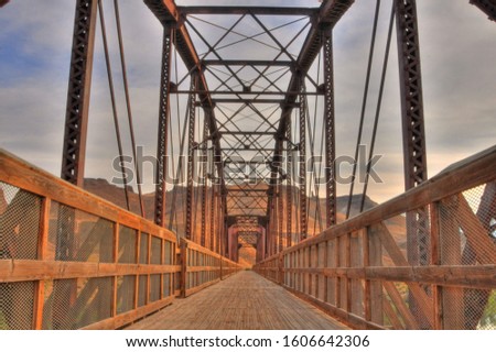 Perspective view a bridge and of its wooden deck walkway with wooden fenced handrails under the metal supporting structures in a warm, rust color