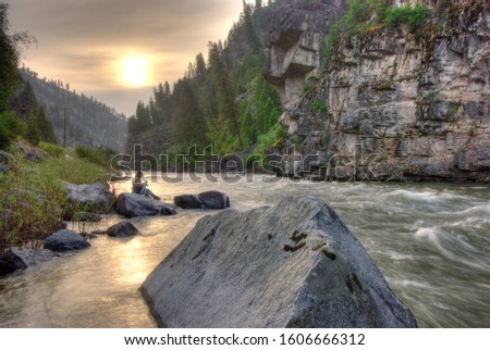 Photographer sitting on a rock on the shore of the Payette River in Idaho checking his camera setting as the Golden Hour approaches