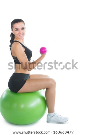 Cheerful active woman sitting on an exercise ball using pink dumbbells smiling at camera
