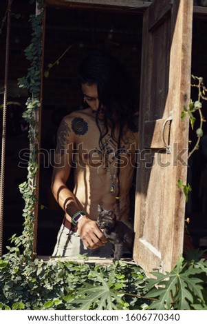 Authentic rural close up outdoor portrait of handsome man with dark skin, tattoo, piercing on fit tan body and  dreadlocks, playing with small black kitten. Human and nature connection concept.