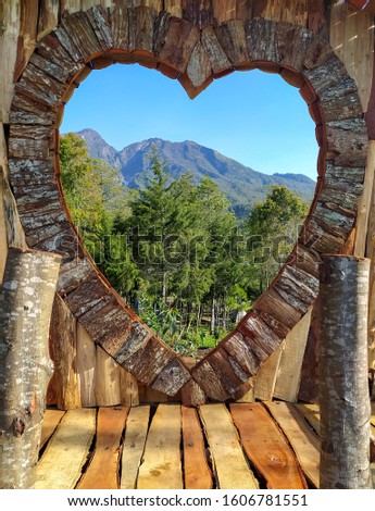 Wood craft is made into a frame shaped as a symbol of love and views of trees, mountains, blue sky in the frame.