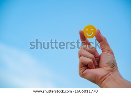 woman hand holding yellow emoticon smiley face icon symbol on blue sky background with copy space, for a positive mindset in business marketing and beautiful world peace concept
