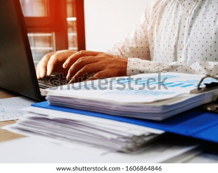 A man works at a computer in the office. Pile of financial performance papers.