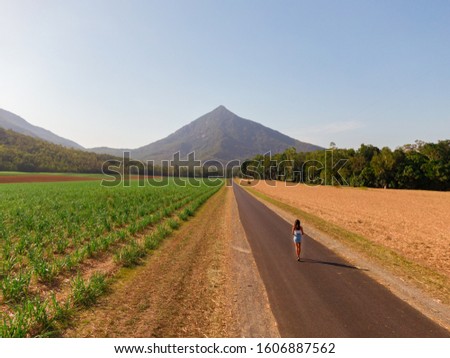Beautiful mountain landscape & Woman with sugar cane fields foreground. Dramatic DRONE aerial view of fields, trees, green forest, farm, mountains & road. Shot in Walsh's Pyramid, Cairns, Australia.