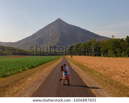 Couple with mountain landscape with sugar cane fields foreground. Dramatic DRONE aerial view of fields, trees, green forest, farm, mountains & road. Romantic shot in Walsh's Pyramid, Cairns, Australia