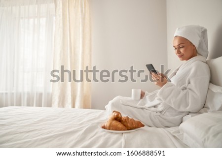 Side view of smiling young woman wrapped in towel and bathrobe waking up in morning using phone on bed. Pretty female drinking coffee and posing in bedroom. Concept of gadgets and social networks.