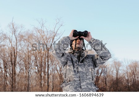 hunter with bristles in military clothes and a blue hat looks through binoculars, the discovery of winter wildlife by man