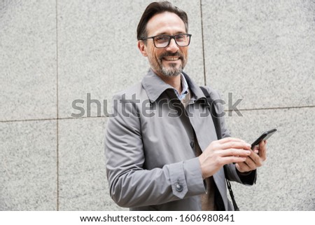 Image of happy adult businessman in eyeglasses using cellphone and smiling while standing near building at city street