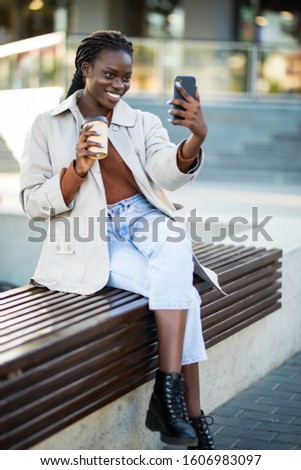 Portrait closeup of friendly african american woman with headphones making video call or take selfie on smartphone while sitting on bench