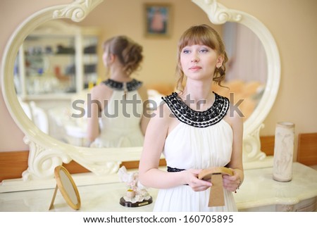 Beautiful dreaming girl stands near mirror and holds frame with photo in light room.