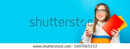 Young woman smiles and holds books and a glass of coffee or tea in her hands on a blue background. Concept of education, college, session, exam, career choice. Banner