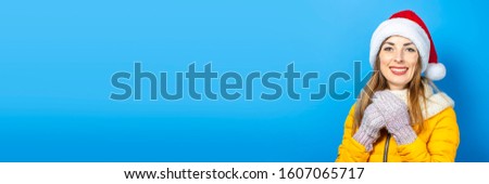 Young woman with a smile in a yellow jacket and hat of Santa Claus on a blue background. Concept of the winter holidays, Christmas, New Year, surprise, shock. Banner