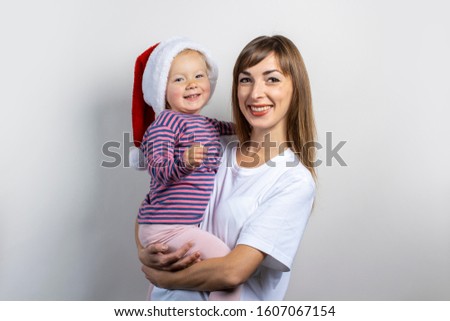 Young woman holds a child in a hat of Santa Claus on a light background. Happy family concept, mom with a child, family holiday, christmas with family