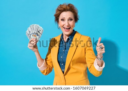 Happy elderly woman holds cash money and like gesture. Photo of business lady in yellow blazer on blue background. Emotions and pleasant feelings concept.