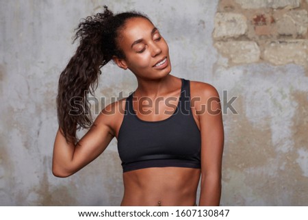 Portrait of young pleased dark skinned woman touching her brown curly long hair with raised hand while posing over brick wall, smiling slightly and keeping eyes closed