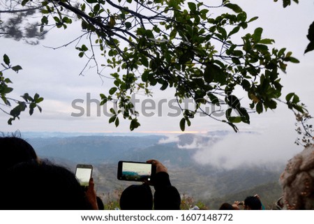 Many people use their phone to take a picture, background has mountain and cloud.