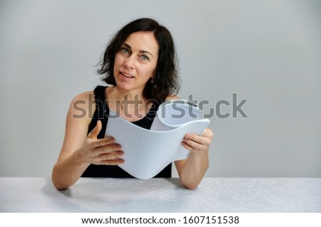 Portrait of a cute smiling talking brunette woman in a black dress on a white background. Sits at a table right in front of the camera with vivid emotions with a folder in his hands.
