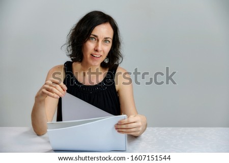 Portrait of a cute smiling talking brunette woman in a black dress on a white background. Sits at a table right in front of the camera with vivid emotions with a folder in his hands.