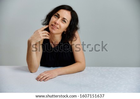Portrait of a cute smiling talking brunette woman in a black dress on a white background. Sits at a table right in front of the camera with vivid emotions.