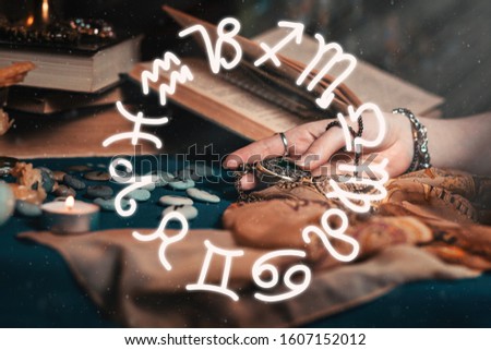 Astrology and esotericism. The female hand of the sorceress holds an amulet in her hands. In the background, old books, fortune-telling runes. In the foreground is the zodiac circle of the horoscope Royalty-Free Stock Photo #1607152012