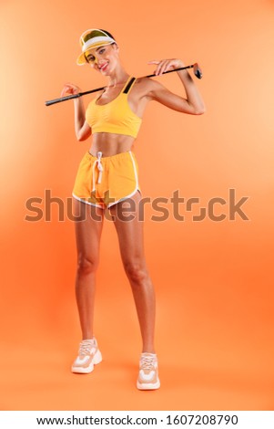 Picture of a cheerful positive young woman posing isolated over yellow wall background in sport top and shorts wearing cap holding golf club.