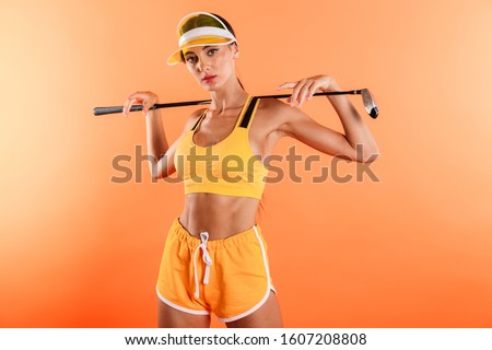 Picture of a serious young woman posing isolated over yellow wall background in sport top and shorts wearing cap holding golf club.