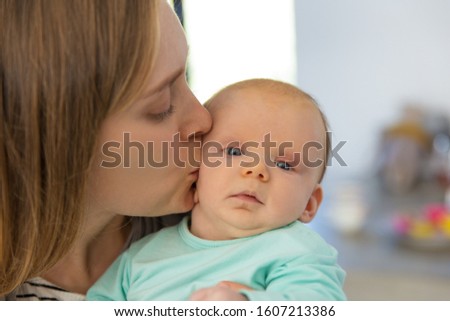 Closeup of new mom holding and kissing baby. Portrait of young woman and cute little child in home interior. Maternity or baby care concept