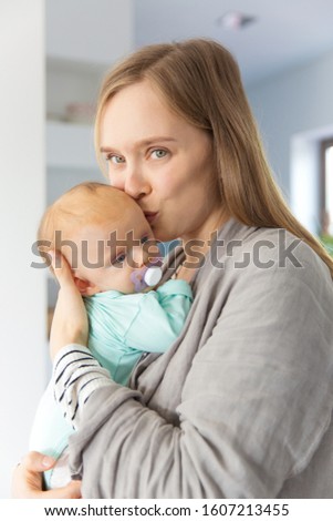 Positive new mother kissing and cuddling baby with soother. Portrait of young woman and cute little child in home interior. Happy new mom concept