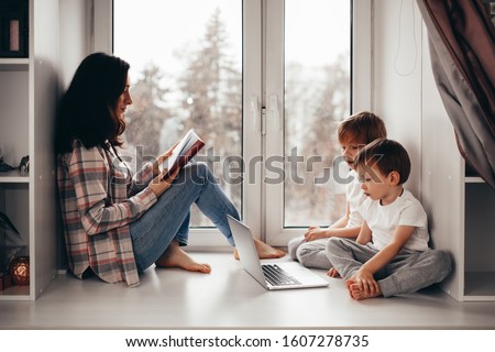 Mom reads a book on the windowsill, children look at the laptop. Authentic life style and technology in childhood.