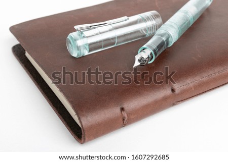 A simple but artful combination of classic leather bound journal and green glass fountain pen set on a plain white background.