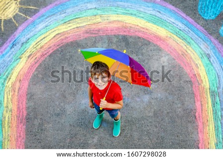 Happy kid boy in rubber boots with rainbow sun and clouds with rain drops painted with colorful chalks on ground or asphalt in summer. Creative leisure for children outdoors in summer