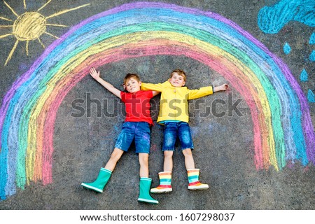 two school kids boys having fun with with rainbow picture drawing with colorful chalks on asphalt. Siblings, twins and best friends in rubber boots painting on ground playing together.