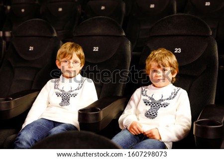 Two little adorable blonde kid boys waiting for a film at cinema. Siblings, twins and brothers having fun with activity of watching movies. Best friends on seats in dark room.