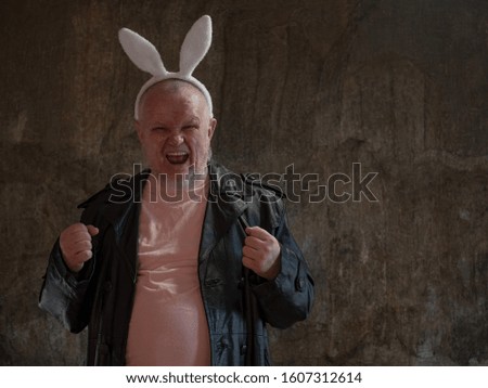 elderly man in pink pajamas, rabbit ears and leather coat of exhibition maniac is dancing against background of imitating mold. concept of perversions of bizarre people. Gray beard and wrinkled face