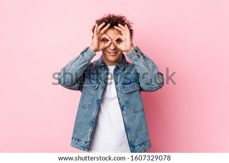 Curly mature man wearing a denim jacket against pink background showing okay sign over eyes
