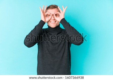 Young caucasian man on a blue background showing okay sign over eyes
