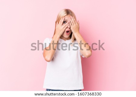 Young blonde woman on pink background blink through fingers frightened and nervous.